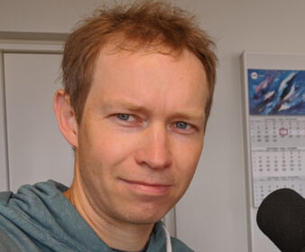 Fotol on Tallinna Tehnikaülikooli teadur Tanel Alumäe. Alumäel on seljas sinakas kampsun, juuksed on veidi sassis ning punakat värvi. Kaadris on näha mikrofoni ning taamal on seina peal kalender.